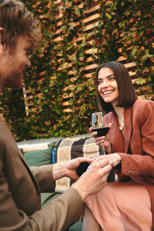 A man in a suit holding a glass of wine next to a woman in a cocktail dress, sharing a moment of...