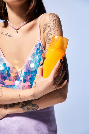A young woman with tattoos, brunette hair, holding a yellow sunscreen in a sunlit studio setting.