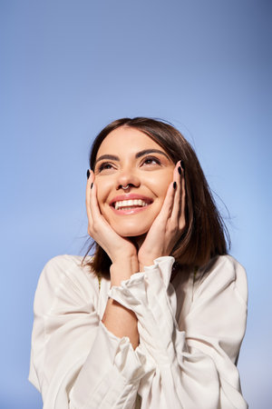A young woman with brunette hair smiles joyfully, hands resting gently on her face in a studio setting.の写真素材