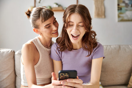 Two women in comfy attire, sitting on a couch, engrossed in a cell phone.