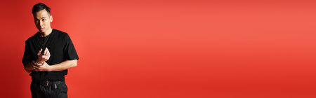 A handsome Asian man stands confidently in front of a striking red wall in a trendy studio setting.