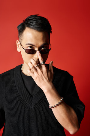 A stylish, handsome Asian man in a black attire holding a sunglasses against a bold red background in a studio setting.の写真素材