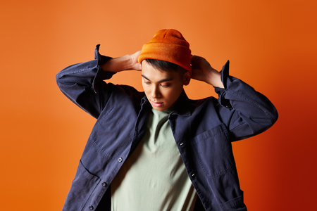 A handsome Asian man, dressed in stylish attire, confidently wears a hat on his head against an orange background.の写真素材