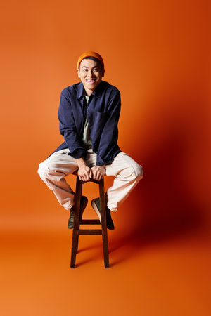 A handsome Asian man in stylish attire sits gracefully on top of a wooden stool against an orange...