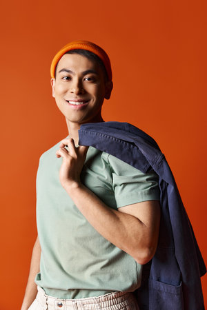 A handsome Asian man in a green shirt is carrying a blue backpack against an orange background.