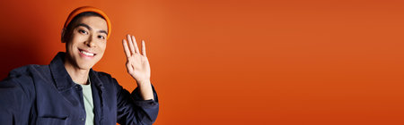 A stylish Asian man in a blue jacket is gesturing with his hand against a vibrant orange background...