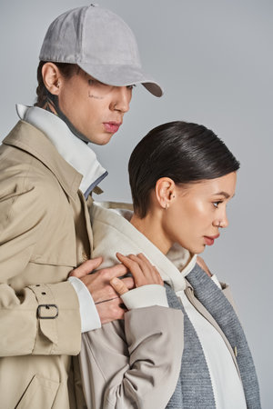 A young stylish couple stands side by side in trench coats in a studio against a grey background.