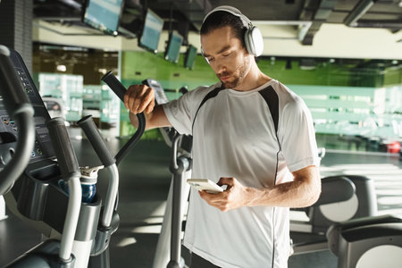 A focused man in athletic wear wearing headphones and using a cell phone while working out in a gym.