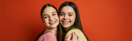 Two young women, pretty and brunette, hug each other affectionately in front of a vibrant red wall.の写真素材