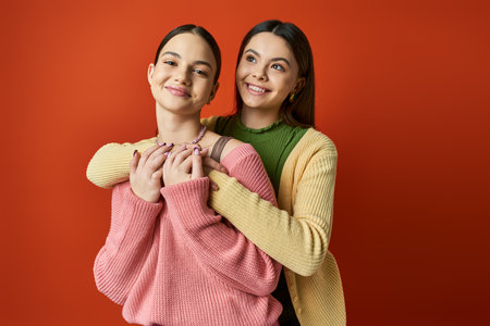 Two pretty, brunette teenage girls hug each other warmly in front of a bold red background,...