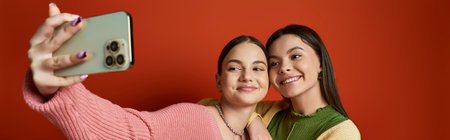 Two young women, friends, taking a selfie with a smartphone against an orange background in a studio setting.の写真素材