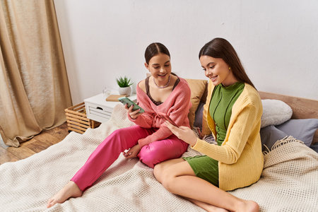 Two teenage girls sit on a bed, engrossed in a cell phone.の写真素材