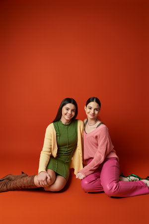 Two pretty, brunette teenage girls sit on the floor in a studio, posing gracefully for a picture against an orange background.の写真素材