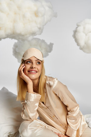A dreamy blonde woman in cozy pajamas sitting atop a white pillow among fluffy clouds.の写真素材