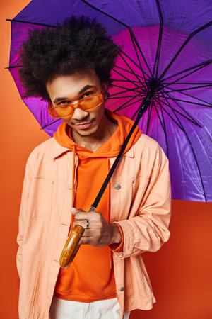 A trendy young African American man with curly hair holding a purple umbrella, exuding emotion on an orange background.の写真素材