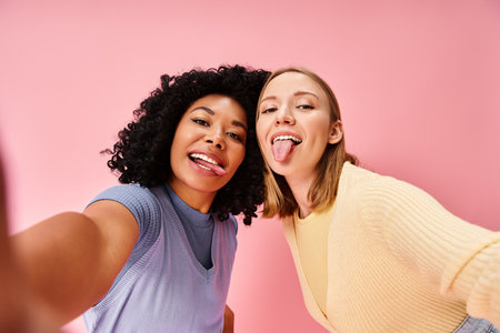 Two women in casual attire taking a selfie in front of a pink wall.