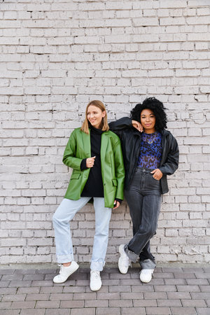 Two attractive diverse women stand near a brick wall.