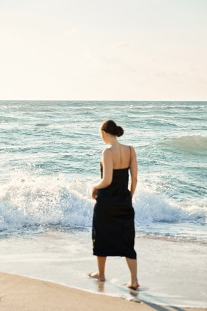 A woman wearing a black dress walks along Miami Beach, Florida, gazing out at the crashing waves.の写真素材