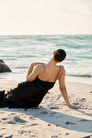 A woman in a black sundress sits on the sand, gazing out at the ocean. The setting sun casts a warm glow on the scene.の写真素材