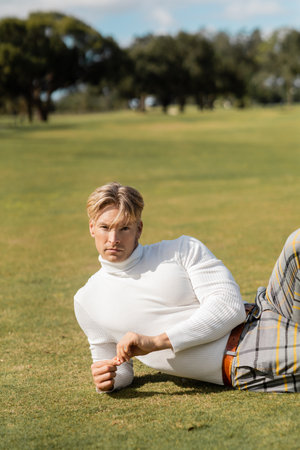 A blonde man in stylish clothing poses on the grass in a Miami park.の写真素材
