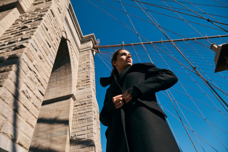 Woman in black coat gazes at sky by Brooklyn Bridge.