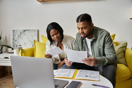An African American couple reviews papers while sitting on a yellow couch in their living room.