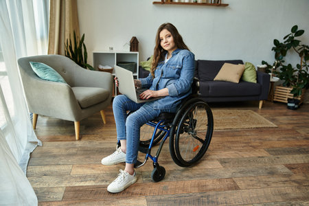 A young woman sits in her wheelchair, smiling, working on her laptop in a modern apartment.