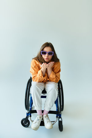Young woman in a wheelchair wears stylish outfit and sunglasses in studio with grey background, sending a kiss with her handsの写真素材