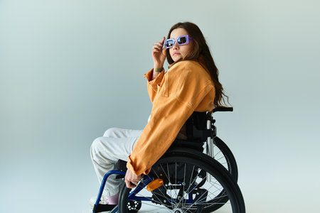 A young woman with long, brunette hair sits in a wheelchair against a grey backdrop, wearing a stylish orange jacket and sunglasses.の写真素材