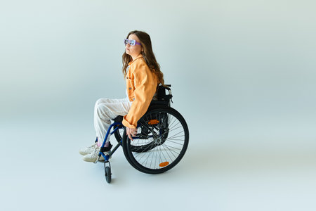 A young woman sits in a wheelchair in a studio with a grey background. She is wearing casual clothing and sunglasses.の写真素材