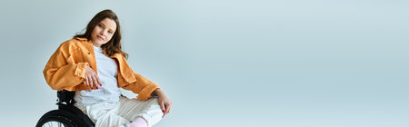 A young brunette woman sits confidently in a wheelchair, dressed in stylish casual attire against a grey background.の写真素材