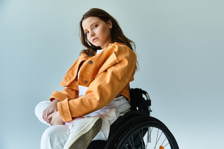 Young woman with long brown hair sits in a wheelchair in a studio, wearing a white shirt and orange jacket against a grey backgroundの写真素材