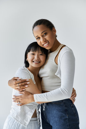 Two young women, one African American and one Asian, share a tender embrace against a simple grey backdrop.の写真素材