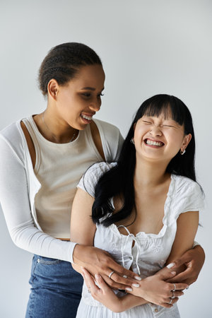 Two young women, one African American and one Asian, stand in a tender embrace, laughing and looking into each others eyes.の写真素材