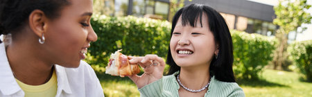 Two young women, one African American and the other Asian, share a laugh and a bite to eat during a picnic.の写真素材