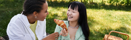 A young African American woman laughs as her Asian partner holds up a croissant during an outdoor picnic.の写真素材