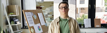 A young man with Down syndrome stands confidently in front of a bulletin board in an office.