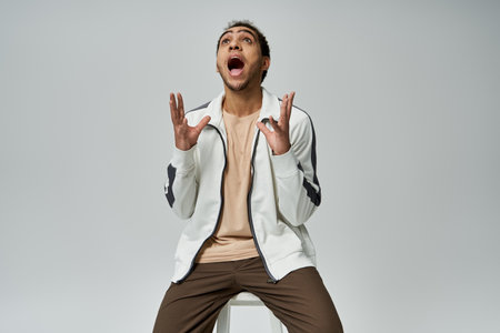 A young African American man is in a stylish white jacket and brown pants against a grey background.