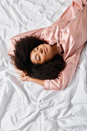 A curly African American woman in pajamas peacefully lays on a white bed in a bedroom. It is morning...