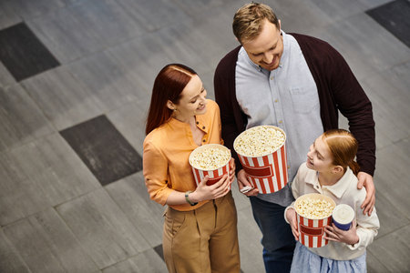 A man and his wife joyfully hold popcorn boxes with kid as they enjoy a movie together at the...