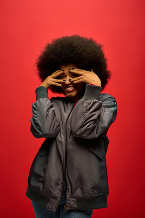 Stylish African American woman in trendy attire covering face with hands.