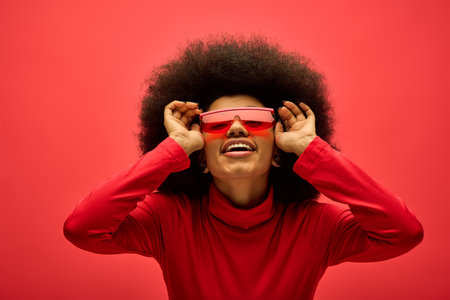Stylish African American woman in red glasses and shirt posing on vibrant backdrop.の写真素材