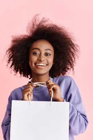 A beautiful young African American woman with curly hair happily holds a shopping bag in a studio...