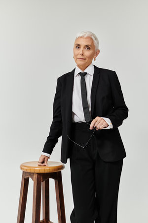 An elegant mature woman stands confidently next to a wooden stool.の写真素材