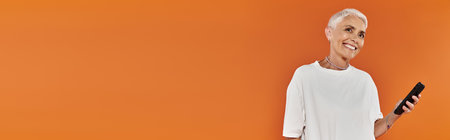 A confident woman smiles while holding her phone against a vibrant orange backdrop.