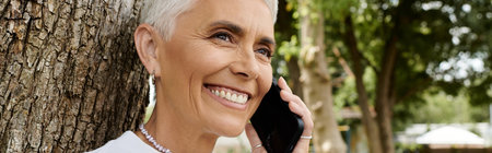A mature woman with short hair smiles while on a phone call outdoors.