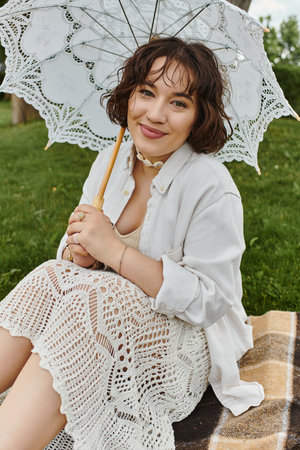 A cheerful young woman relaxes on a blanket, savoring a sunny summer picnic with a delicate umbrella.の写真素材
