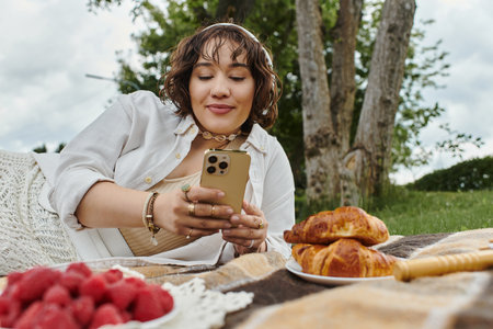 A young woman in a white shirt enjoys a summer picnic, relaxing with her phone and delicious pastries.の写真素材