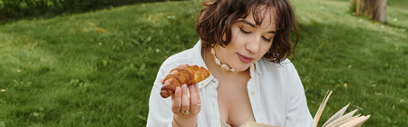 A young woman in a white shirt relaxes on a summer day, savoring a croissant while reading a book.の写真素材