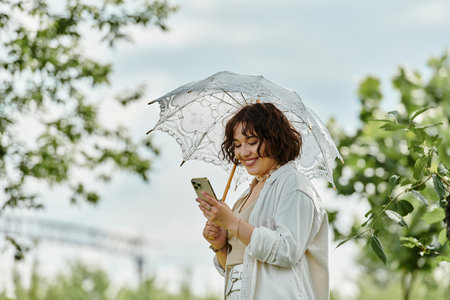 A cheerful woman wears a white shirt, delighting in her phone while shaded by an elegant umbrella in summer greenery.の写真素材